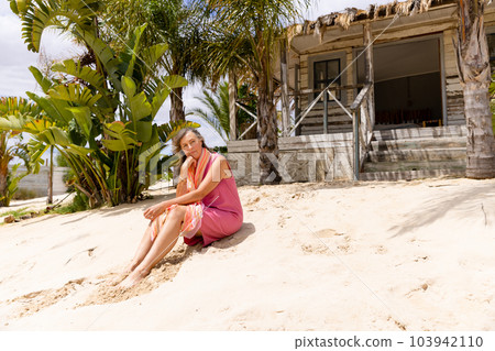 Thoughtful caucasian senior woman sitting on sandy beach against wooden cottage on sunny day Thoughtful caucasian senior woman sitting on sandy beach against wooden cottage on sunny day 103942110