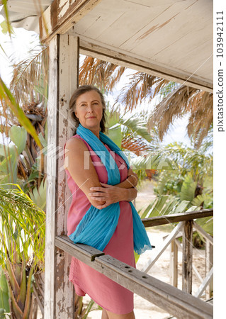 Caucasian pensive senior woman with scarf and arms crossed standing by railing in balcony Caucasian pensive senior woman with scarf and arms crossed standing by railing in balcony 103942111