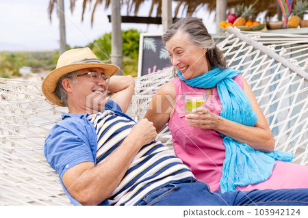 Caucasian senior woman holding drink and lying with husband on hammock at beach 103942124