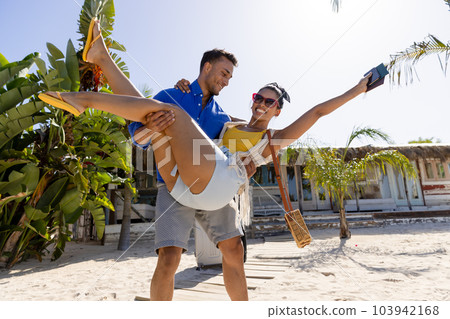 Caucasian young boyfriend carrying excited girlfriend while standing at beach against clear sky 103942168