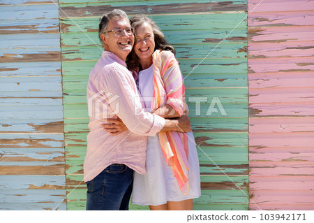 Cheerful caucasian senior couple embracing and looking away while standing against wooden wall 103942171