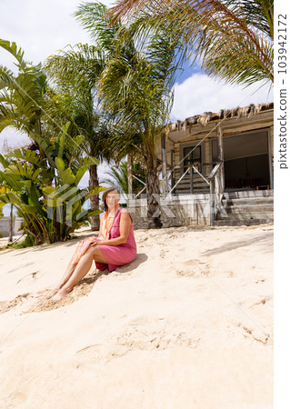 Caucasian senior woman sitting on sandy beach against palm trees and cottage during sunny day Caucasian senior woman sitting on sandy beach against palm trees and cottage during sunny day 103942172