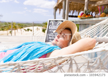 Caucasian senior woman wearing hat sleeping on hammock at beach during sunny day 103942173