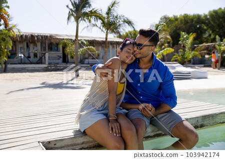 Happy caucasian young man hugging girlfriend while sitting at poolside at tourist resort against sky 103942174