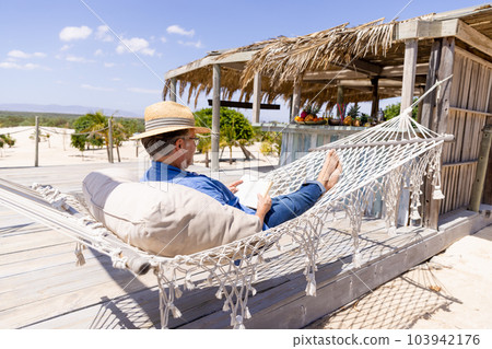 Caucasian senior man wearing hat and reading book while lying on hammock at beach against sky Caucasian senior man wearing hat and reading book while lying on hammock at beach against sky 103942176