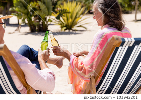 Happy caucasian senior couple toasting beer bottles while sitting on deck chairs at beach 103942201