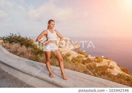Fitness woman sea. A happy middle aged woman in white sportswear exercises morning outdoors in a park with a beach view. Female fitness pilates yoga routine concept. Healthy lifestyle. 103942546