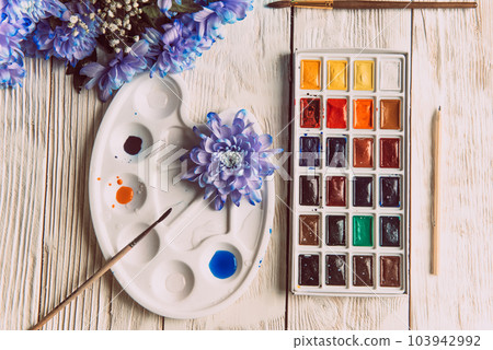 Palette of watercolor paints, brushes and paper for a water color on wooden background, close up. Flat lay, spring background with copy space Palette of watercolor paints, brushes and paper for a water color on wooden background, close up. Flat lay, spring background with copy space 103942992