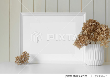 Empty picture frame with beautiful bouquet of dried hydrangea flowers. Modern white ceramic vase with dry grass. Wooden wall background. Scandinavian Empty picture frame with beautiful bouquet of dried hydrangea flowers. Modern white ceramic vase with dry grass. Wooden wall background. Scandinavian 103943626