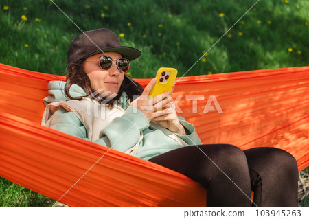 A woman in a cap lies in a hammock and looks at her phone A woman in a cap lies in a hammock and looks at her phone 103945263