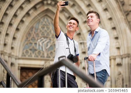 Two men in shorts with luggage doing selfie Two men in shorts with luggage doing selfie 103945465