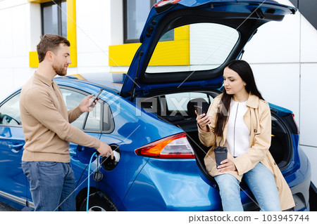 Young couple man and woman traveling by electric car having stop at charging station. Boyfriend plugging in cable to charge. Man talking with girlfriend, holding cup drinking hot coffee smiling 103945744