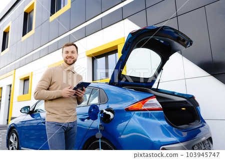 A young man is charging an electric car while standing at a station and holding a tablet 103945747
