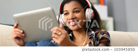 Relaxed charming african american woman in headphones listening to music and choosing with finger on tablet screen Relaxed charming african american woman in headphones listening to music and choosing with finger on tablet screen 103945850