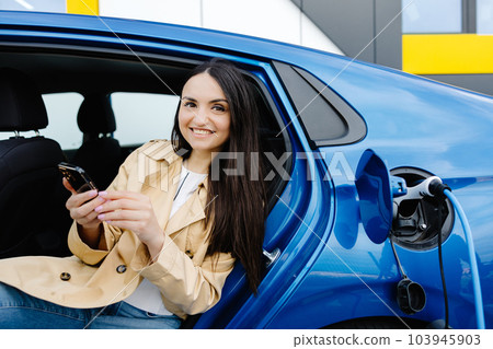 Business lady sitting inside charging electric car and using modern smartphone. Technology and transport concept. Business lady sitting inside charging electric car and using modern smartphone. Technology and transport concept. 103945903