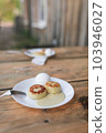 Two white plates with cheesecakes, sweet sauce, eggs and metal fork on aged wooden table. Village atmosphere and early morning breakfast for two persons, with old village house background. 103946027