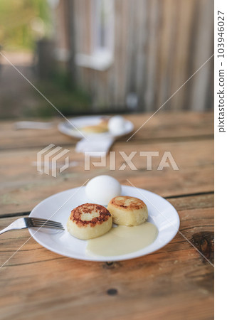 Two white plates with cheesecakes, sweet sauce, eggs and metal fork on aged wooden table. Village atmosphere and early morning breakfast for two persons, with old village house background. Two white plates with cheesecakes, sweet sauce, eggs and metal fork on aged wooden table. Village atmosphere and early morning breakfast for two persons, with old village house background. 103946027