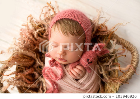 A cute little newborn baby in a white winding and a red cap sleeps sweetly with a red knitted bunny in small hands. Wicker basket. Professional macro photo against a light wooden floor. 103946926
