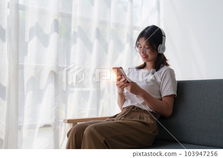 Young relaxed smiling pretty woman relaxing sitting on sofa at home. Female feeling joy enjoying with mobile phone on cozy couch 103947256