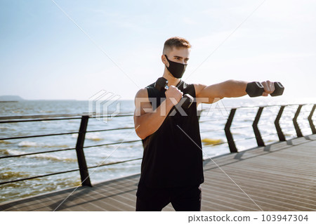 Handsome muscular man in protective mask, sports outfit doing functional exercises on seaside promenade. 103947304