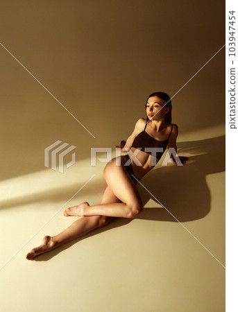 Sensual, beautiful young brunette girl with slim body sitting on floor, posing in black underwear against studio background with warm sunlight Sensual, beautiful young brunette girl with slim body sitting on floor, posing in black underwear against studio background with warm sunlight 103947454