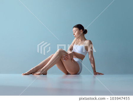 Self-care. Beautiful young girl with well-kept skin and slim body, sitting on floor, posing in underwear against blue studio background Self-care. Beautiful young girl with well-kept skin and slim body, sitting on floor, posing in underwear against blue studio background 103947455