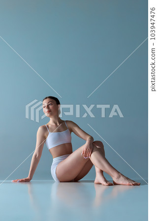 Portrait of tender, pretty young girl with slim body, sitting on floor, posing in underwear against blue studio background 103947476