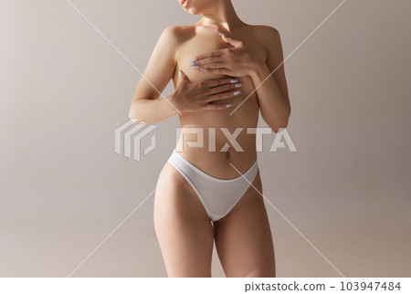 Cropped image of young woman with slim body covering breast with hand. Model posing in underwear against grey studio background. Mammoplasty Cropped image of young woman with slim body covering breast with hand. Model posing in underwear against grey studio background. Mammoplasty 103947484