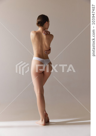 Full-length rear view of tender girl with slim body, healthy back, buttocks. Model posing in underwear against grey studio background 103947487