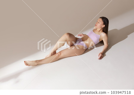 Top view young girl with fit, slim figure lying on floor, posing in underwear against grey studio background. Tenderness 103947518