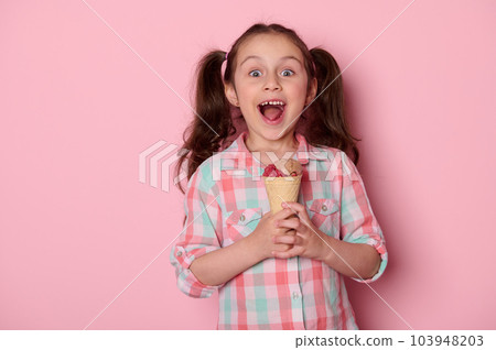 Studio portrait on pink background of a beautiful mischievous Caucasian little kid girl with a tasty ice cream in waffle cone, expressing happiness, surprise and positive emotions, looking at camera Studio portrait on pink background of a beautiful mischievous Caucasian little kid girl with a tasty ice cream in waffle cone, expressing happiness, surprise and positive emotions, looking at camera 103948203