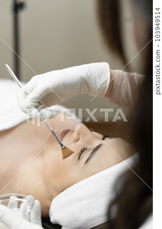 The photo reveals the uniqueness and skill of the cosmetologist during facial skin peeling, which turns your skin into youth. Professional cosmetologist applies a mask for cleaning and peeling The photo reveals the uniqueness and skill of the cosmetologist during facial skin peeling, which turns your skin into youth. Professional cosmetologist applies a mask for cleaning and peeling 103949514