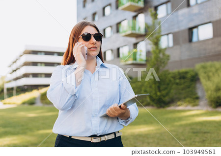 Lifestyle portrait of a stylish young businesswoman talking on cell phone, making call and working online, standing on city street. Successful business. Happy female using modern technology Lifestyle portrait of a stylish young businesswoman talking on cell phone, making call and working online, standing on city street. Successful business. Happy female using modern technology 103949561