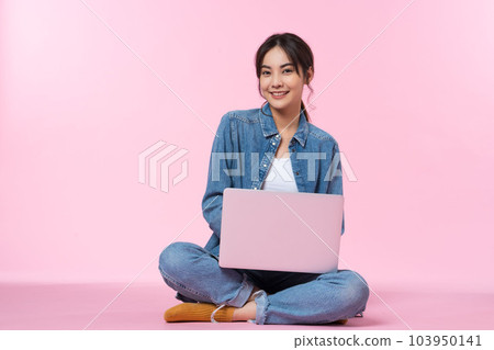 Young Asian college student sits cross-legged on floor working with a laptop computer, looking directly at the camera in a studio shot on a pink background 103950141