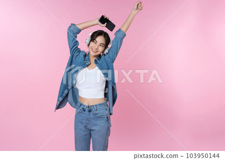 Young Asian woman wearing headphones dancing and listening to music from smartphone shoot in isolated on pink background 103950144