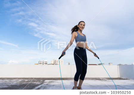 Young Asian woman with jump rope on rooftop. Fitness female doing skipping workout outdoors 103950250