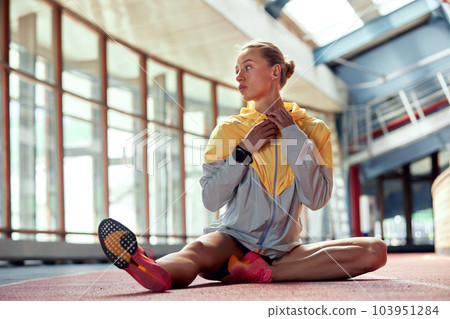 A young athlete girl is warming up before the race at the stadium. A slender blonde in a black tracksuit and a gray-yellow jacket sits on the ground and stretches her muscles before a competition. 103951284