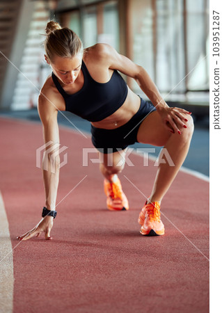 A young athlete girl is warming up before the race at the stadium. A slender blonde in a black tracksuit stretches her leg muscles before a competition on the track. Sports and recreation. 103951287