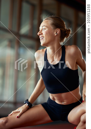 A young athlete girl is warming up before the race at the stadium. A slender blonde in a black tracksuit is concentrating before the competition, sitting on the track in a relaxed pose. A young athlete girl is warming up before the race at the stadium. A slender blonde in a black tracksuit is concentrating before the competition, sitting on the track in a relaxed pose. 103951288