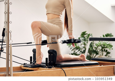 A young girl is doing Pilates on a reformer bed in a bright studio. A beautiful slender brunette in a beige bodysuit does exercises to strengthen the muscles of her arms and shoulder girdle. A young girl is doing Pilates on a reformer bed in a bright studio. A beautiful slender brunette in a beige bodysuit does exercises to strengthen the muscles of her arms and shoulder girdle. 103951349