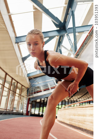 Young athlete girl is warming up before the race at the stadium. A slender blonde in a black tracksuit stretches her leg muscles before training. Sports and recreation. 103951393