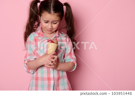 Adorable happy little child girl with two ponytails, wearing checkered shirt, holding a waffle cone with delicious ice cream over isolated pink background. Food. Summer 103951448