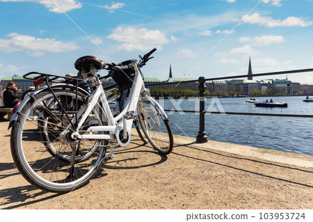 Scenic view two modern bicycles parked at embankment of Alster lake in Hamburg street old city center background summer day panorama view. Healthy travel lifestyle concept. Urban rental transport 103953724