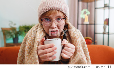 Sick woman girl wear hat wrapped in plaid sit alone shivering from cold on sofa drinking hot tea 103954911