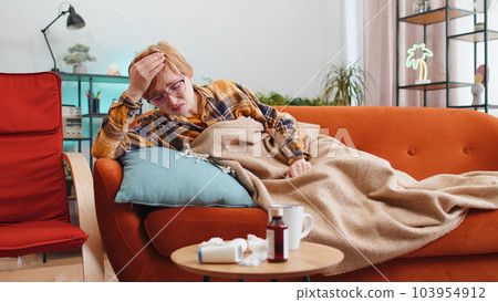 Sick ill woman suffering from cold or allergy sitting on home sofa sneezes wipes snot into napkin Sick ill woman suffering from cold or allergy sitting on home sofa sneezes wipes snot into napkin 103954912