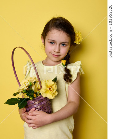 Beauty portrait Caucasian noble charming little girl in yellow elegant wear and flowers in hair, holding bouquet of yellow roses in purple wicker basket, smiling looking at camera, isolated background Beauty portrait Caucasian noble charming little girl in yellow elegant wear and flowers in hair, holding bouquet of yellow roses in purple wicker basket, smiling looking at camera, isolated background 103955526