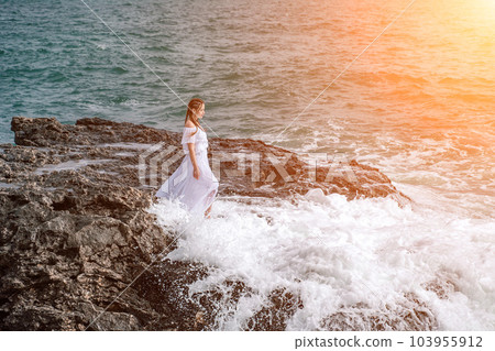 A woman stands on a rock in the sea during a storm. Dressed in a 103955912