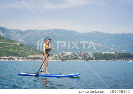 Young women Having Fun Stand Up Paddling in blue water sea in Montenegro. Against the backdrop of the Montenegrin mountains. SUP Young women Having Fun Stand Up Paddling in blue water sea in Montenegro. Against the backdrop of the Montenegrin mountains. SUP 103956163
