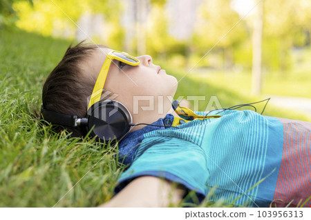 Happy boy in sunglasses lying on the grass and listening to music in headphones 103956313