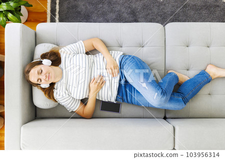 Woman in headphones laying, relaxing on a comfortable couch, listening to music 103956314